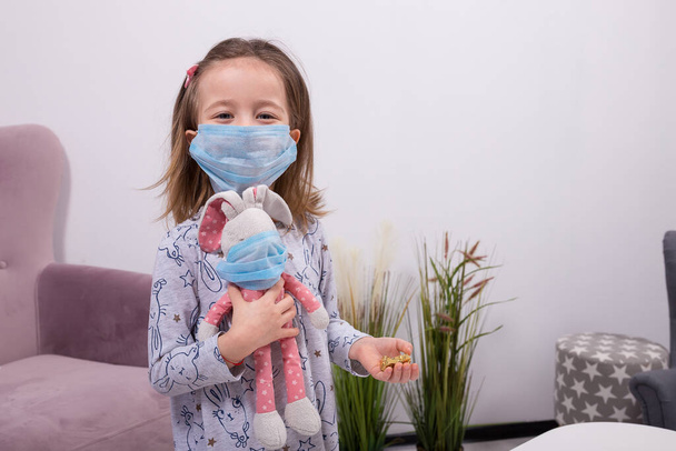 funny little girl with a protective mask holding in her arms a rabbit doll and some chocolates. Quarantine at home. - Photo, Image