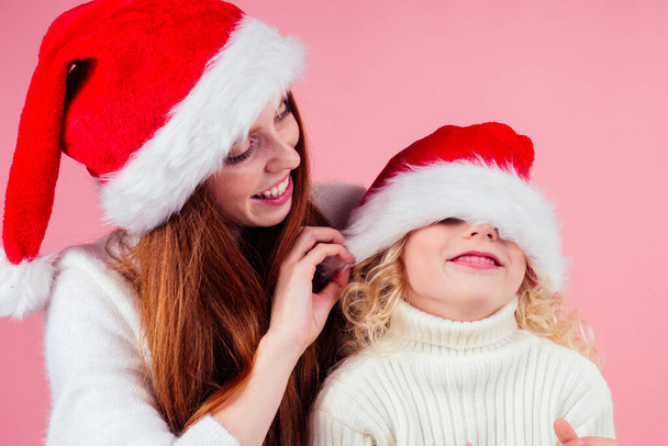 ,happy redhead ginger woman and cute little blonde girl hiding eyes under santa claus hat and having fun together in studio pink background - Fotoğraf, Görsel