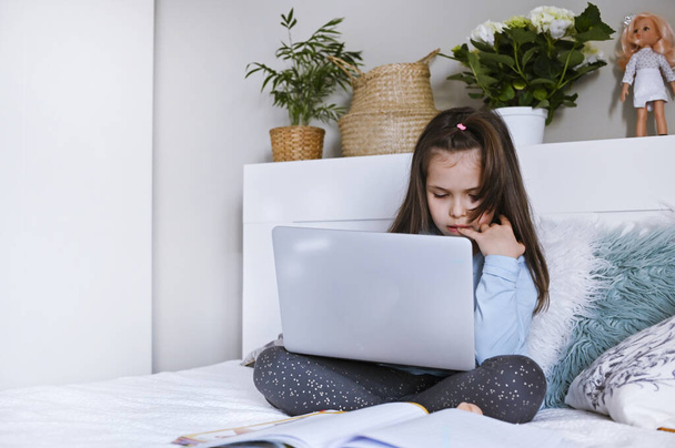 Pretty schoolgirl studying homework her online lesson at home, social distance during quarantine, self-isolation, online education concept. Girl with school books and a laptop on the bed in the room. - Φωτογραφία, εικόνα