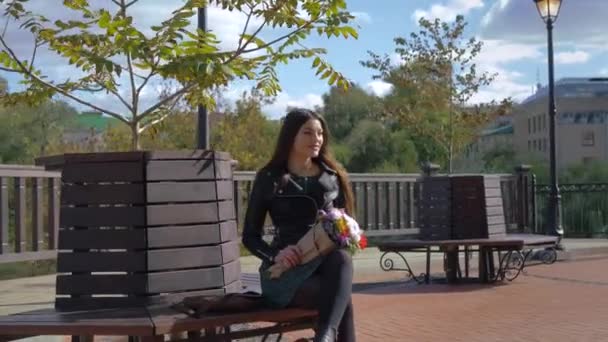 girl with a bouquet of flowers is sitting on a wooden bench under tree. Waiting for her lover in the city center - Imágenes, Vídeo