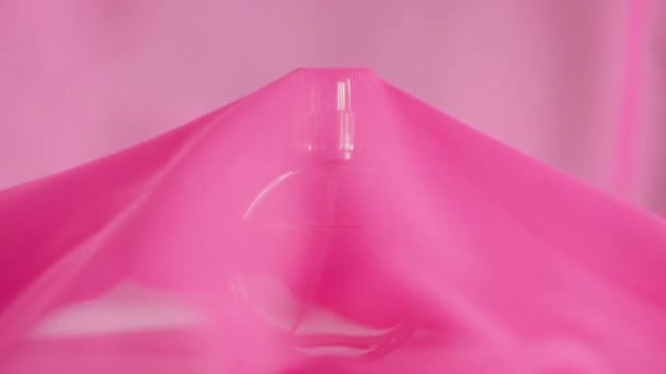 Oval surfaced bottle with pink perfumes or essential oils is on the table. Pink fabric flutters around and waves in the air and around the bottle. Concept of aroma and smell. Close up. Slow motion - Footage, Video