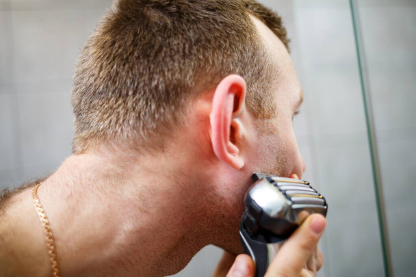 Um homem rapa a cara com uma navalha eléctrica à frente de um espelho. Irritação da pele. Procedimento de banho - Foto, Imagem