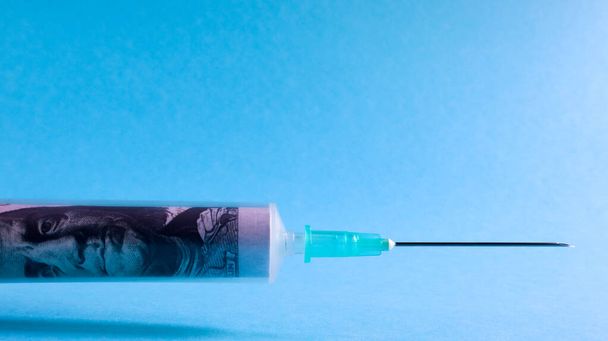 A roll of 100 dollars money inserted into a white medical hypodermic needle on a blue background. The concept of health care costs, credit, investment. Copy space for your text or logo - Fotografie, Obrázek
