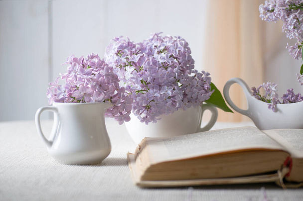 Room interior with lilacs flower in vase and old vintage open fairytale book on table and curtain hanging on wall, tender romantic spring home decor in morning light, soft focus. - Fotó, kép