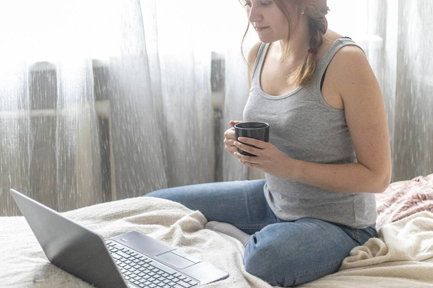 Young beautiful woman working on a laptop in the morning lying on bed at home. Cute girl with cup of coffee or tea doing shopping online on website. Remote work in quarantine on self-isolation. - 写真・画像