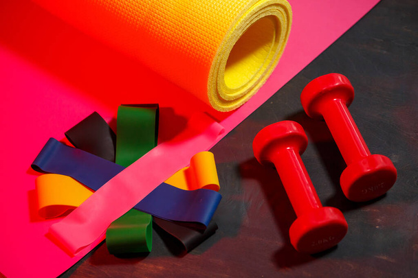 Red dumbbells, fitness mat and elastic bands for sports on a pink background. Healthy lifestyle. Fitness equipment for weight training. Muscle development and fitness training - Photo, Image