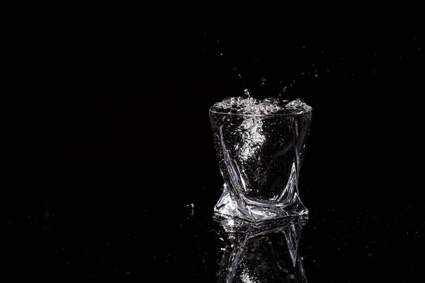 Black background is a glass in which a drop of water falls. Splashing water on glass - Foto, imagen