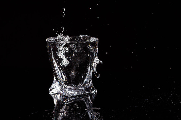 Black background is a glass in which a drop of water falls. Splashing water on glass - Photo, Image