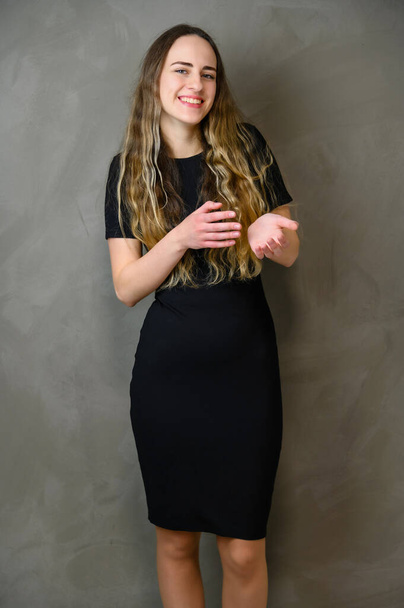 Vertical photo of a slender young Caucasian girl model with long curly hair in a black dress on a gray background. Made in the studio. - Фото, зображення