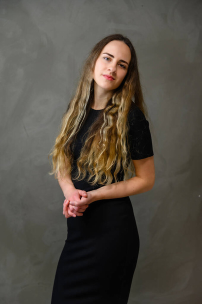 Vertical photo of a slender young Caucasian girl model with long curly hair in a black dress on a gray background. Made in the studio. - Photo, image