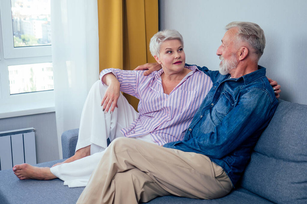 Mature good-looking well dressed couple sitting in sofa in day light from big window and yellow curtains in living room,touching hand - Photo, Image