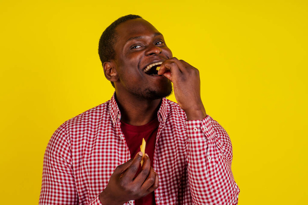 handsome latin man eating fries with appetite and enjoyment in studio yellow background - Foto, Bild