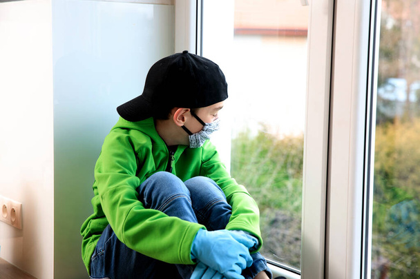 Хлопець носить медичну маску. Дитина сидить біля вікна і дивиться на вулицю. Він виглядає нудним і сумним. Він може захворіти або карантирувати через Коронавірус 19 - Фото, зображення
