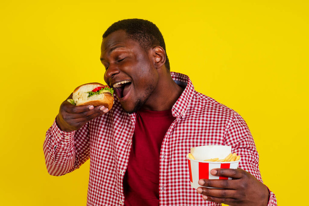 handsome latin man eating fries nuggets and burger with appetite and enjoyment in studio yellow background.chicken legs in a bucket - Photo, image