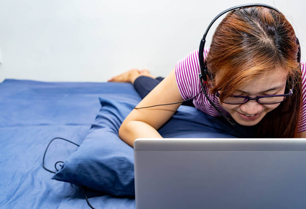 Ένα κορίτσι που εργάζεται από το σπίτι στο κρεβάτι της με φορητό υπολογιστή και ακουστικά. Μαμά που εργάζονται από το σπίτι κατά τη διάρκεια της παραμονής ειδοποίηση στο σπίτι λόγω της επιδημίας του ιού - Φωτογραφία, εικόνα