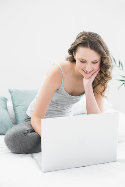 Smiling casual brunette looking at laptop in bed - Фото, изображение