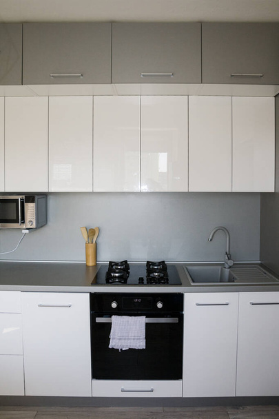 北欧、ミニマリズムの家。ホワイトキッチンデザイン。シルバーのアクセントとアクセサリーと現代的な白い光沢のあるキッチン。居心地の良い、美しい、機能的なキッチンデザイン. - 写真・画像