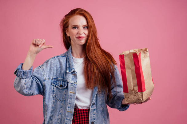 frowning long red-haired ginger woman holding paper eco bag package with chips in studio pink background.she doesnt want to eat unhealthy food - Fotografie, Obrázek