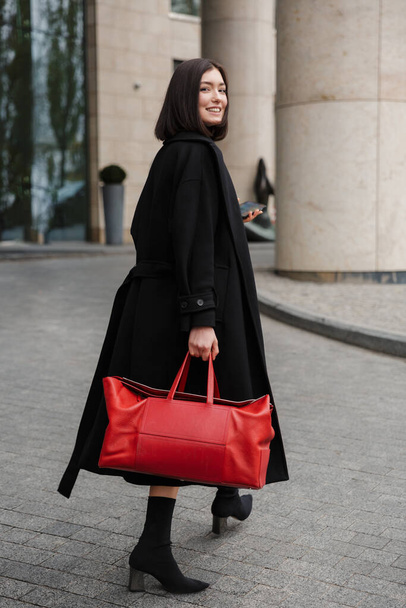 Attractive young brunette woman wearing autumn coat walking in a street, carrying travel bag - Fotografie, Obrázek