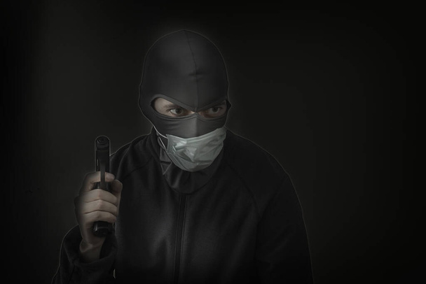 Bandit in einer schwarzen Maske mit einem Schlitz für die Augen und einer medizinischen Maske mit einer Waffe in den Händen. Niedrige Luke. Nahaufnahme. Kopierraum - Foto, Bild