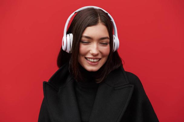 Beautiful smiling young brunette woman wearing autumn coat standing isolated over pink background, listening to music with wireless headphones - Foto, afbeelding