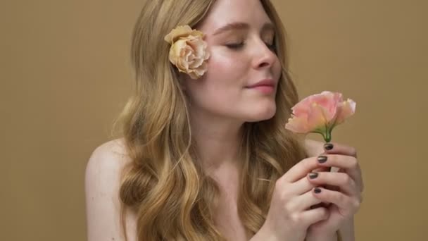 A gorgeous young half-naked woman with long hair is feeling the smell of a beautiful flower isolated over beige background - Filmati, video