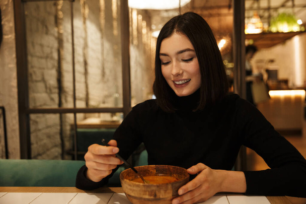 Beautiful young brunette woman sitting at the cafe table indoors, eating soup - Foto, afbeelding