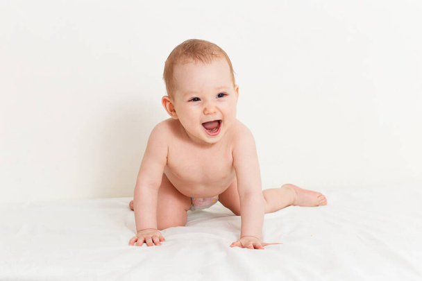 Eight-month-old toddler in diaper crawls on bed on white sheet - Fotoğraf, Görsel