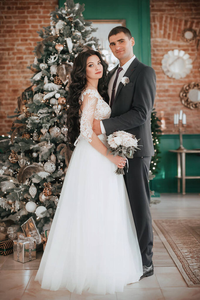 Krásná a mladá nevěsta a ženich pózují v blízkosti vánočního stromečku - Fotografie, Obrázek