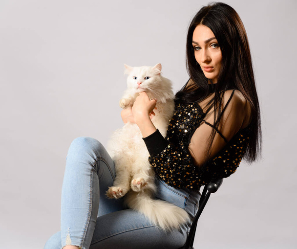 Portrait of caucasian brunette girl model on a white background in studio. Model is sitting on a chair with a white cat in her arms. - Foto, afbeelding