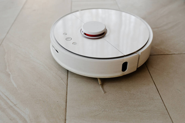 Robot porszívó tisztítás közben, fehér robot porszívó takarítja a padlót a törmeléktől, otthoni tisztítás elektromos porszívóval, porszívó elektromos robot tisztító technológia - Fotó, kép