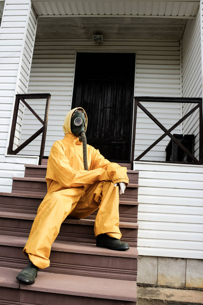 Una mujer con una máscara de gas con tubo flexible de respiración de goma y un mono protector amarillo sentado en las escaleras de la casa. Contaminación ambiental y autoaislamiento, concepto de cuarentena
 - Foto, Imagen