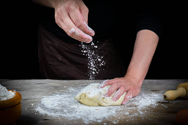 Ο άνθρωπος χέρια ζυμώνει ζύμη ψωμιού σε ένα ξύλινο τραπέζι με μαύρο φόντο. Έννοια του σκάφους - Φωτογραφία, εικόνα