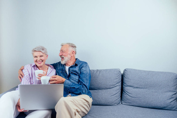 elderly couple in love sitting gray couch in living room look at screen laptop and drinking cup of tea or coffee copyspase - Fotografie, Obrázek