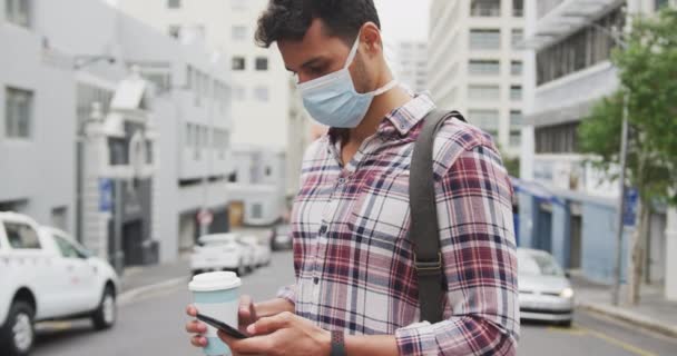 Retrato de um homem caucasiano usando uma máscara facial contra coronavírus, vívido 19 para fora e sobre nas ruas da cidade durante o dia, usando seu smartphone
. - Filmagem, Vídeo
