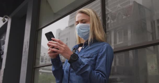 Caucasian woman wearing a face mask against coronavirus, covid 19 out and about in the city streets during the day, using her smartphone. - Záběry, video