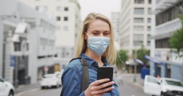 Caucasian woman wearing a face mask against coronavirus, covid 19 out and about in the city streets during the day, using her smartphone and looking straight into a camera. - Filmagem, Vídeo