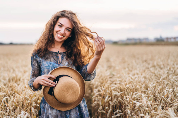 Closeup portrait of a beautiful young woman with curly hair. Woman in dress and hat standing in wheat field. - Fotografie, Obrázek