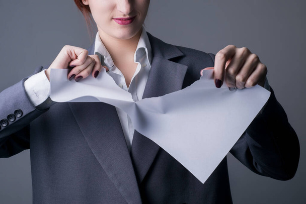 close-up. Studio portrait of a young business woman, with red lipstick, and a gray business jacket, tearing a blank paper, with a grin. On a gray background. Promotional photo. Office Style. Girl boss breaks the contract, grimacing, angry mouth. Tear - Foto, imagen