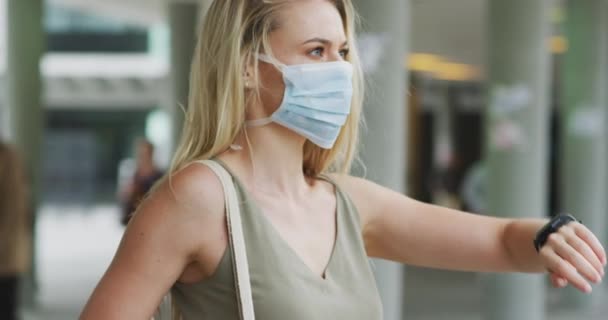 Caucasian woman wearing a face mask against coronavirus, covid 19 out and about in the city streets during the day, using a smartwatch, while a man is passing by in the background. - Filmati, video