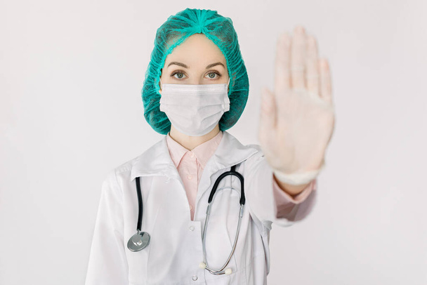 Coronavirus pandemic, infectious diseases outbreak. Young woman doctor infectionist in medical mask and cap showing stop gesture by hand, as symbol of keep distance, avoid communication, stay at home - Foto, afbeelding