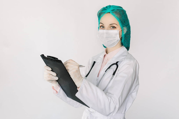 Portrait of young pretty female doctor wearing white coat, medical cap and face mask, holding clipboard and writing notes, looking at camera, standing on isolated white background - Fotó, kép