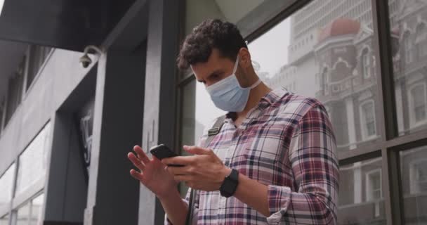 Portrait of a Caucasian man wearing a face mask against coronavirus, covid 19 out and about in the city streets during the day, using his smartphone. - Кадры, видео