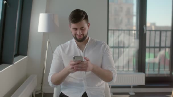 Smiling young man in white shirt is chatting in social networks using smartphone - Séquence, vidéo