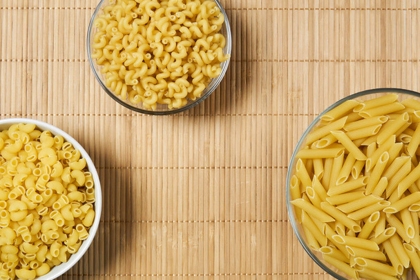 three bowls with dry short pasta on a cane tablecloth, background, top view - Photo, image
