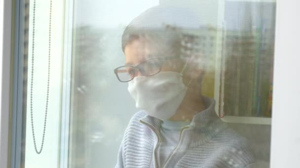Egy fiatal fiú orvosi maszkban és szemüvegben kinézett az ablakon. Önizolálás karanténban, koronavírus, kovid 19. - Felvétel, videó