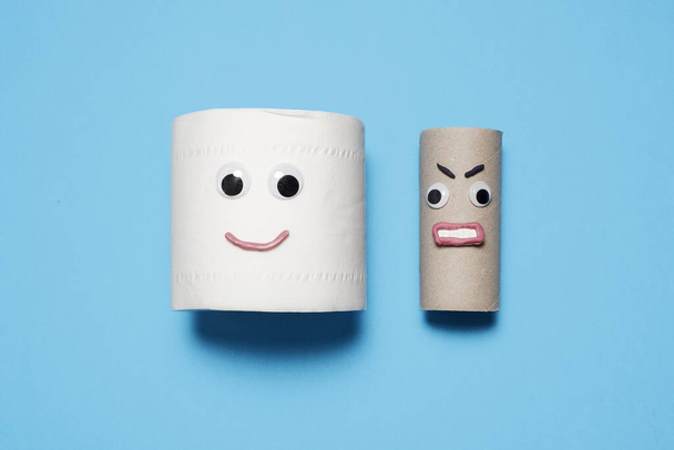 Glücklich lächelnd eine ganze Rolle Toilettenpapier neben einer wütenden leeren Rolle mit googlen Augen und Mund auf blauem Hintergrund mit Kopierraum und Platz für Text. - Foto, Bild