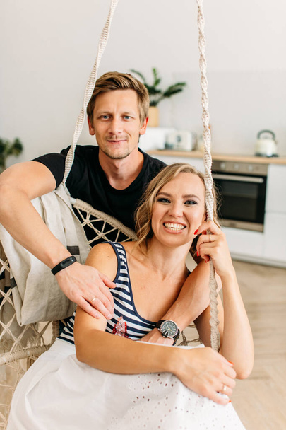 young beautiful couple, a girl in a white skirt and a striped T-shirt, a guy in a dark T-shirt, they are sitting in a bright room, a man hugs a girl from the back, she is very funny and laughs - Foto, Bild
