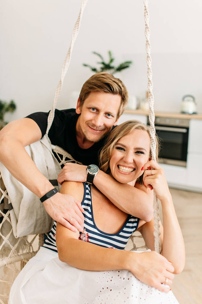 young beautiful couple, a girl in a white skirt and a striped T-shirt, a guy in a dark T-shirt, they are sitting in a bright room, a man hugs a girl from the back, she is very funny and laughs - Foto, Imagen