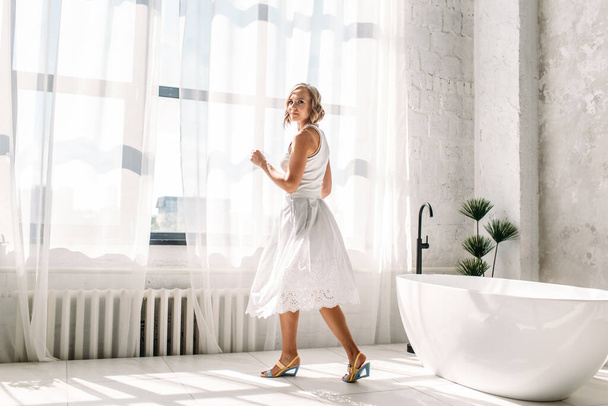 young beautiful girl in a white skirt and white t-shirt, in a spacious bright bathroom, they are in the rays of the sun from a large window, behind it is a white bathtub, she is happy, has a rest, laughs, smiles - Foto, Bild